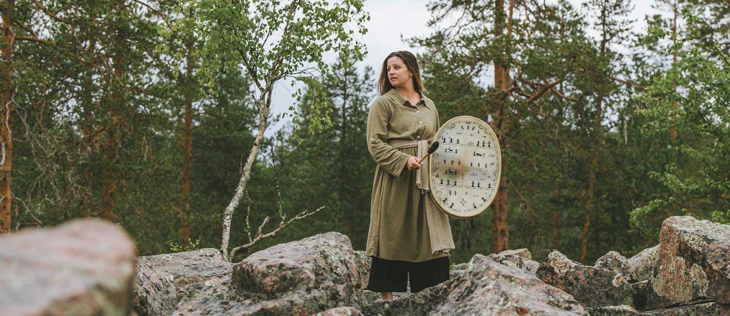 Nainen soittaa lapinrumpua mäntymetsässä kalliolla